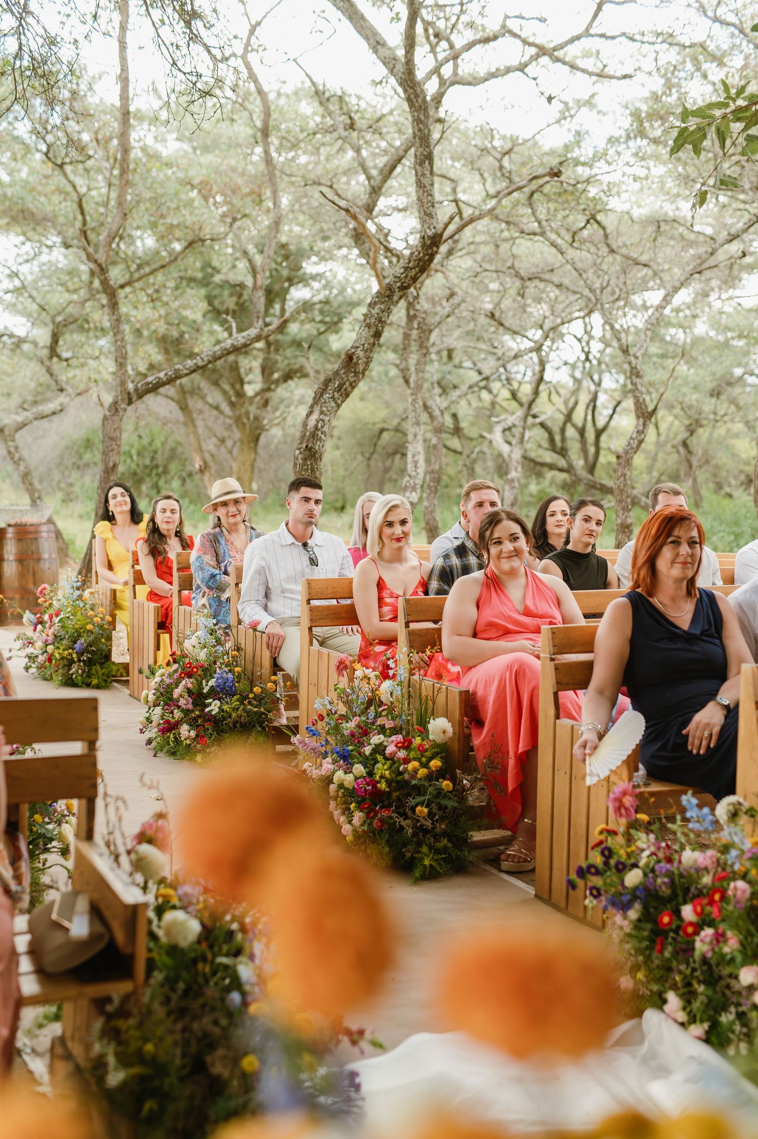 Wedding guests seated on wooden pews along flower-lined aisle at colorful outdoor ceremony surrounded by trees.