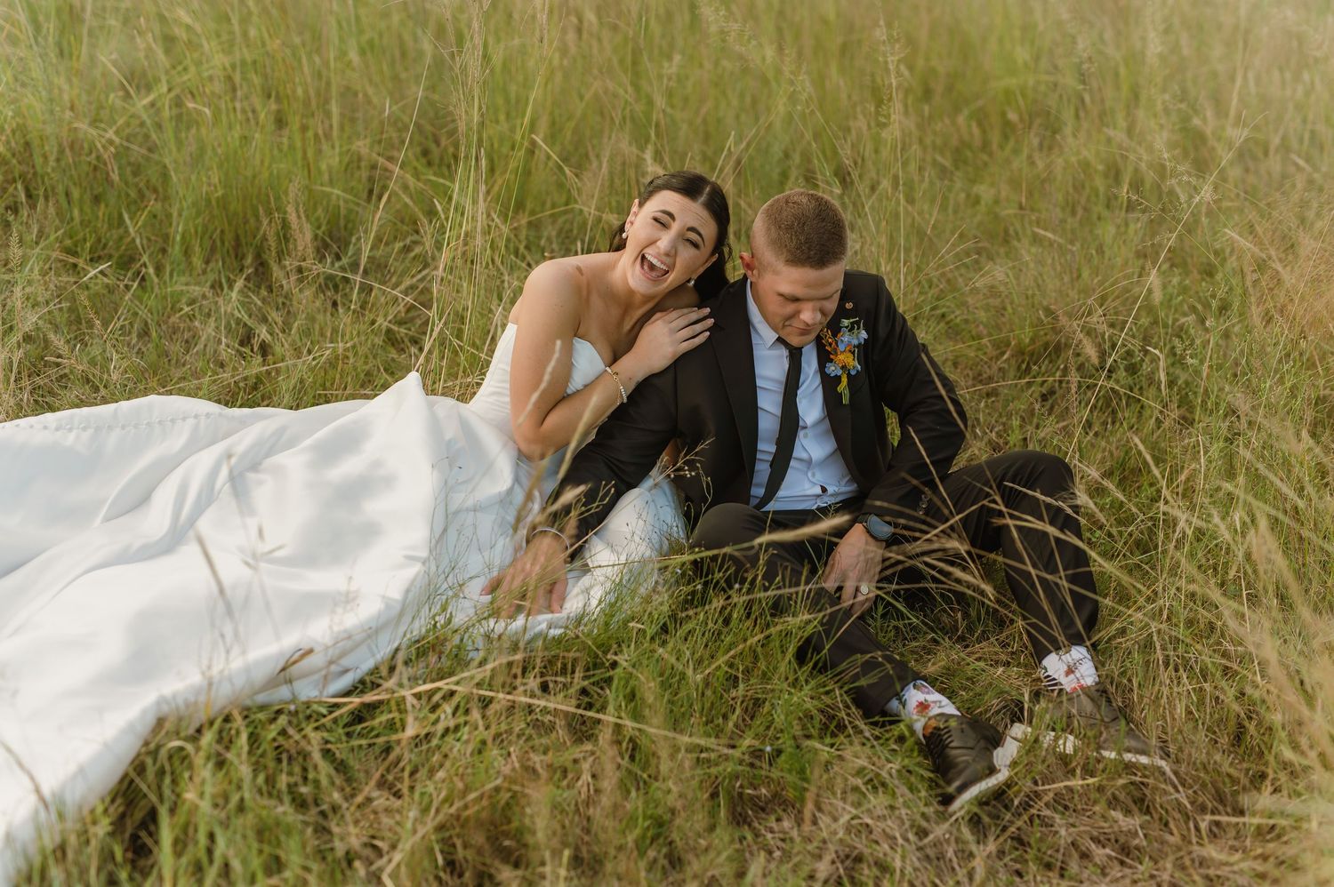 Laughing bride and groom sitting together in tall golden grass during a relaxed outdoor portrait session.