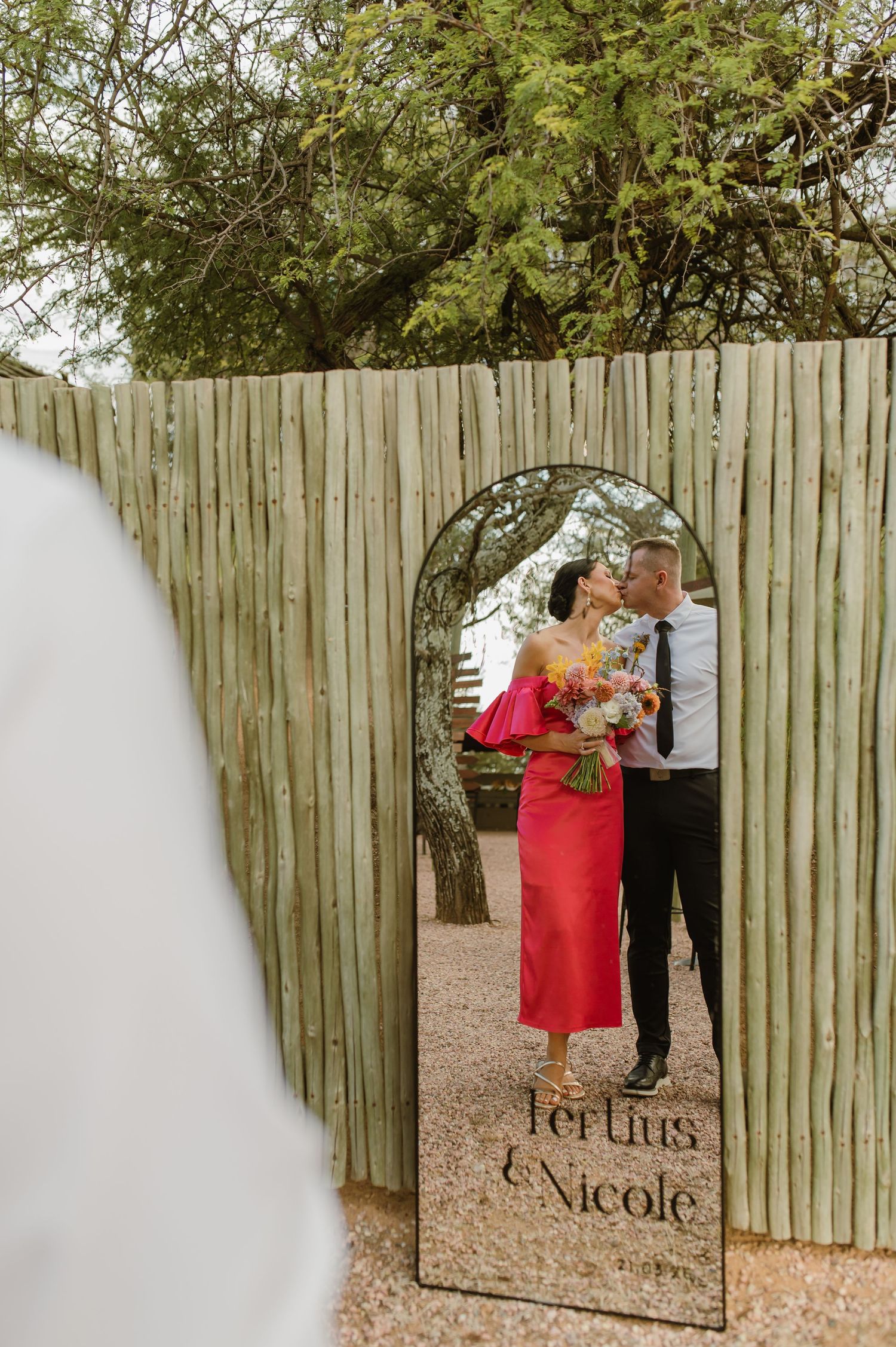 Bride and groom kissing reflected in arched mirror engraved with Tertius and Nicole at outdoor venue.