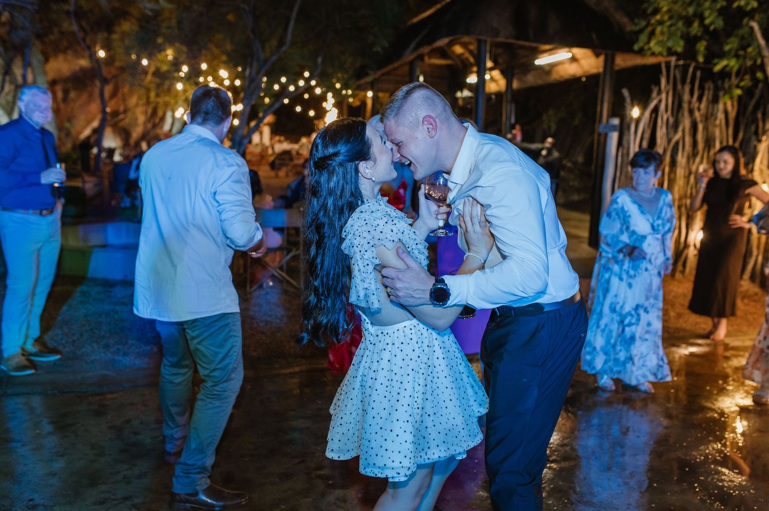 Couple sharing romantic kiss while dancing outdoors at night under string lights at wedding reception.