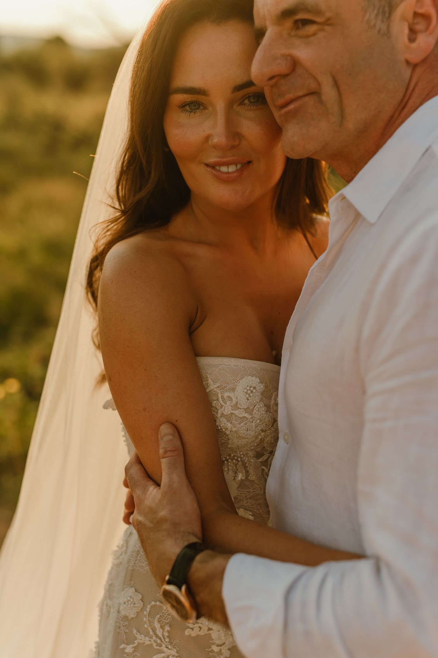 Bride in lace strapless gown embraces groom in white shirt during golden hour outdoor wedding portrait.