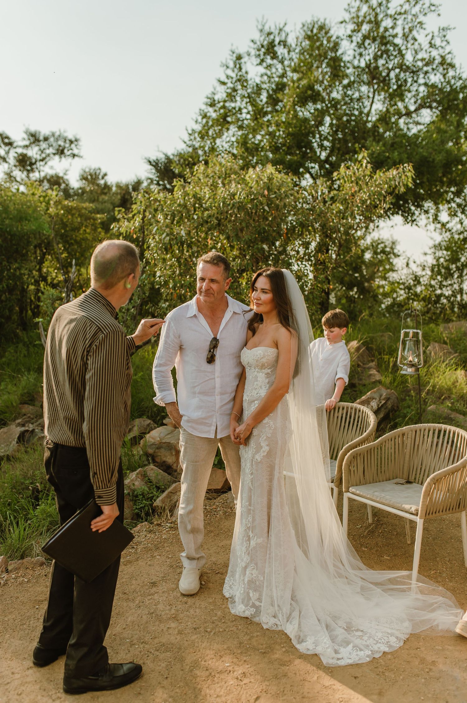 Outdoor wedding ceremony with bride in lace gown and veil, groom in white shirt, and officiant in lush garden setting.