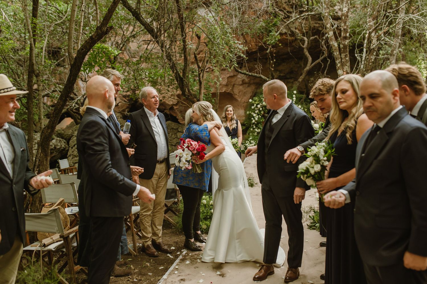 Wedding guests line a forest pathway during an outdoor ceremony surrounded by lush greenery.