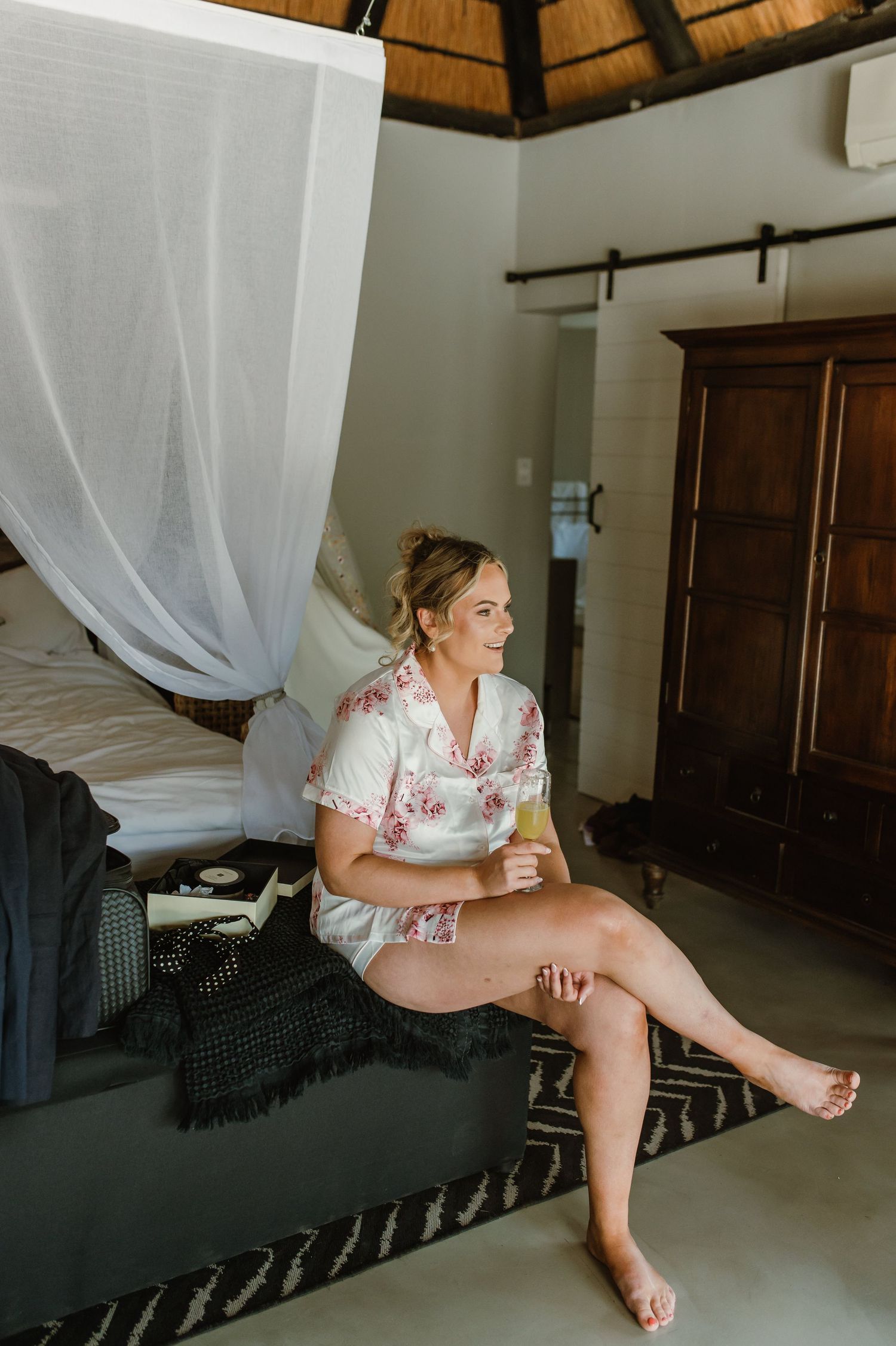 A person in a floral robe relaxes on a lounge chair in a rustic room with a thatched roof.