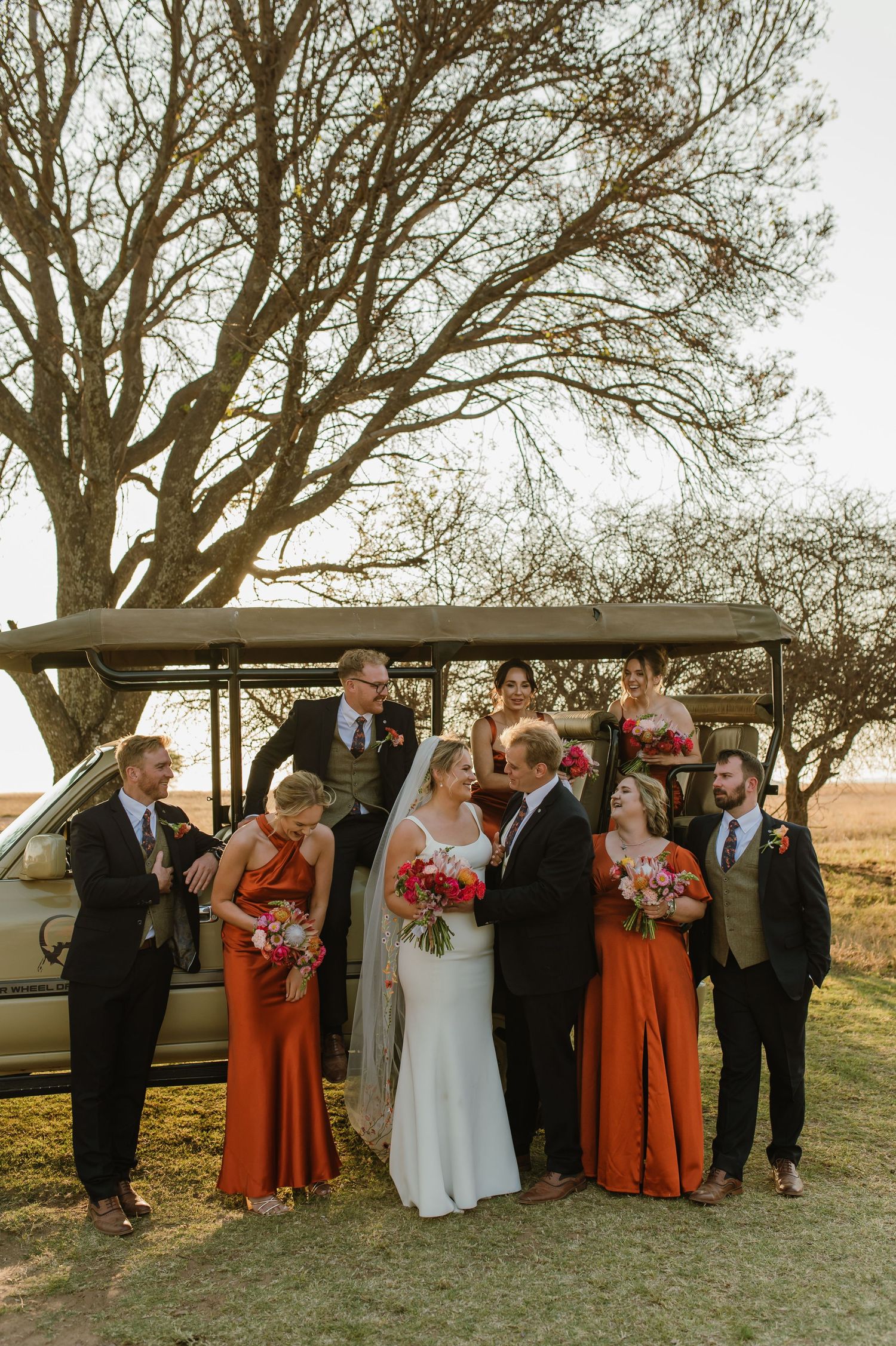 Wedding party gathered for formal photos beside a classic car under sprawling trees.
