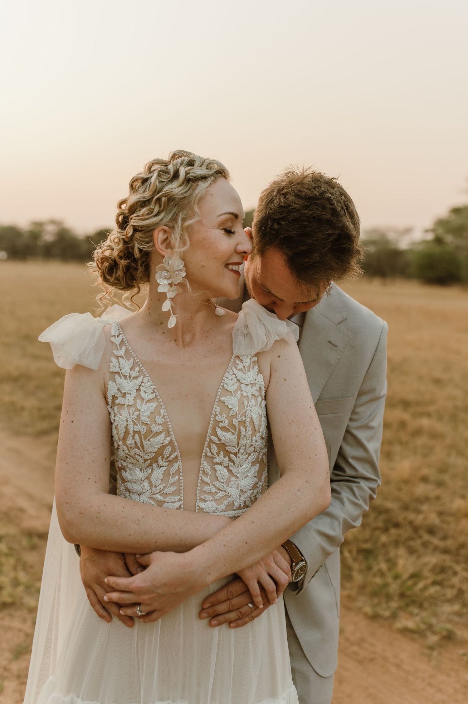 Romantic wedding couple embraces in a sun-drenched field wearing elegant bridal attire.