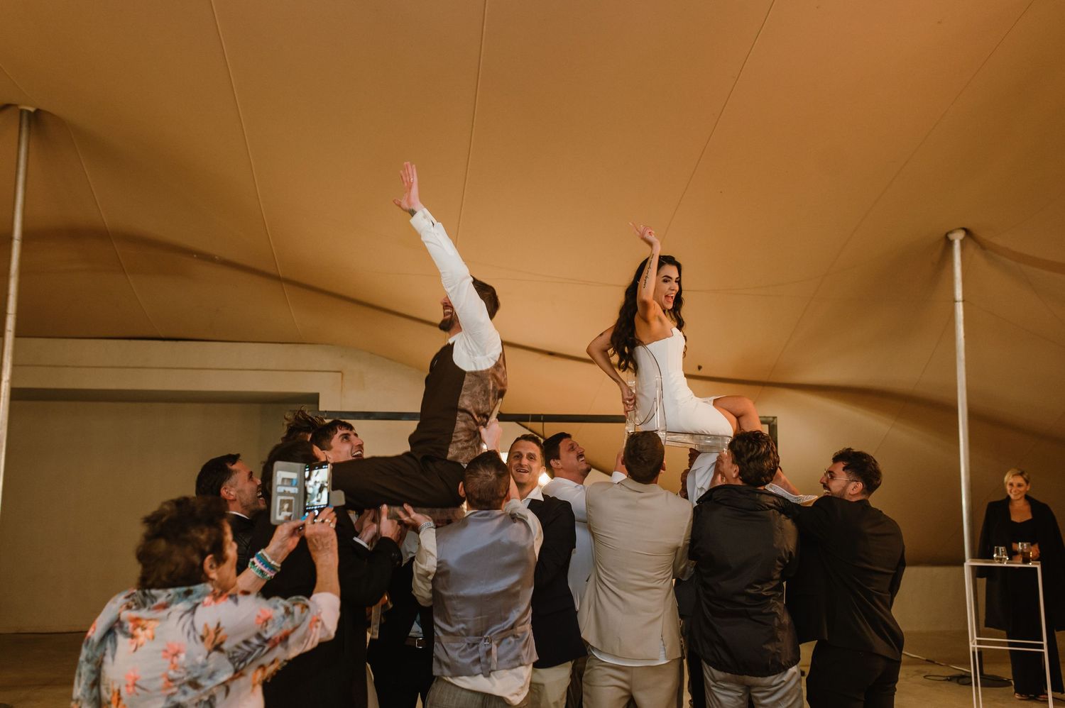 Wedding guests lift and celebrate with the couple during a lively reception under a tent with string lights.