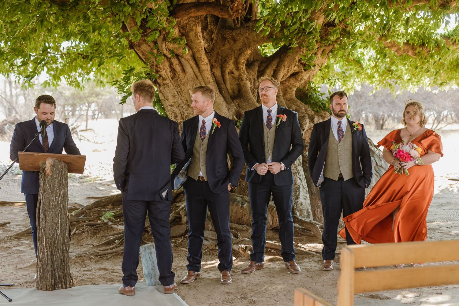 Wedding guests gather under trees for an outdoor ceremony with wooden benches.