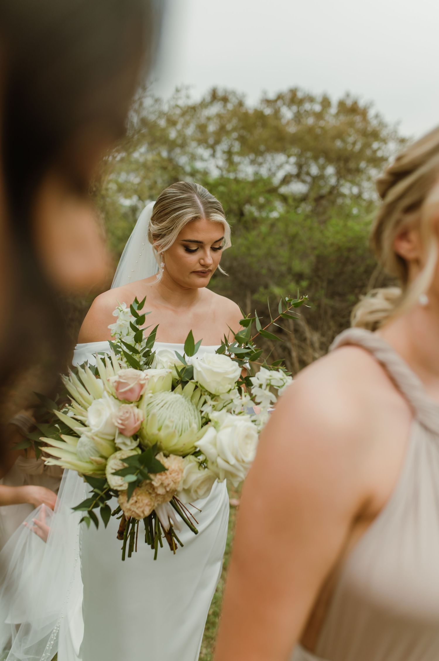 Bride admires her white and blush floral bouquet while surrounded by bridesmaids.