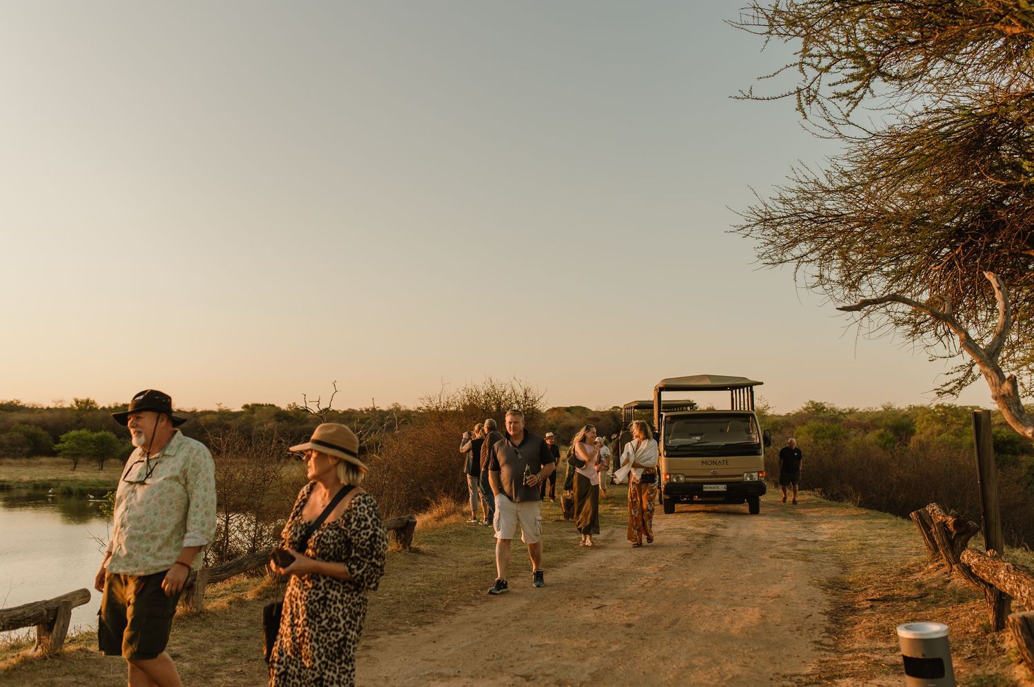 Safari tour group walking along dirt path at sunset with game vehicle in African wilderness.