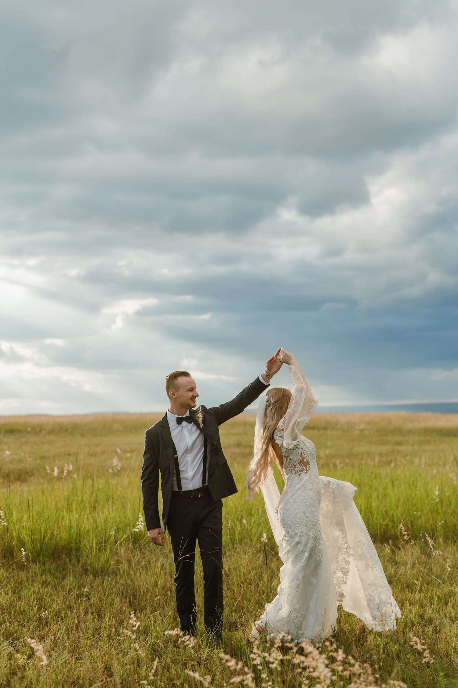 Newlyweds dancing playfully in a golden meadow with stormy skies overhead.