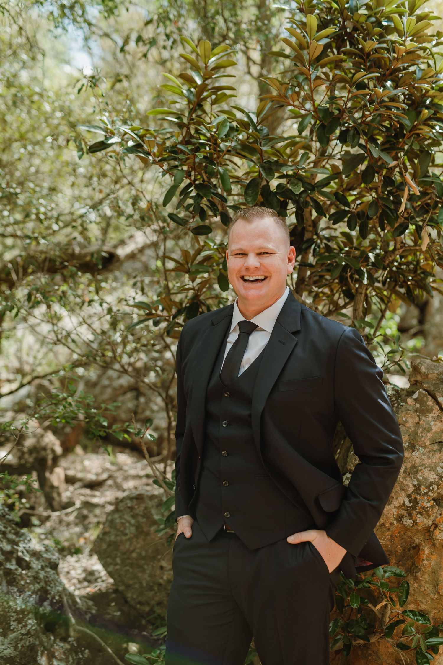 Wedding party member in black suit smiling in an outdoor garden setting.
