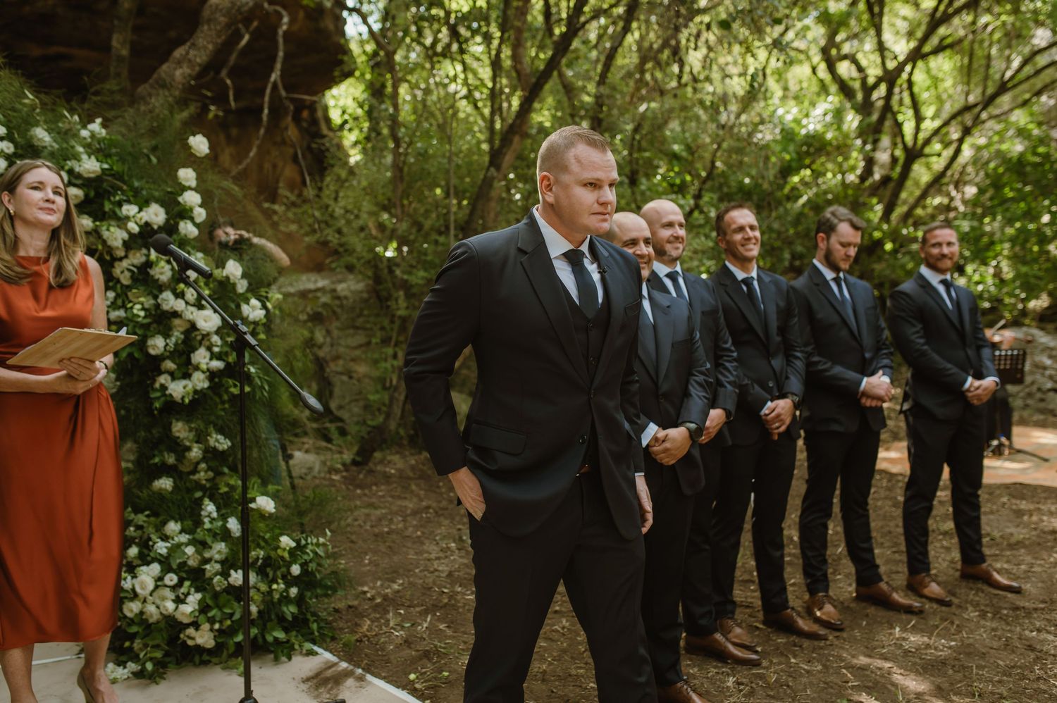 Wedding party members positioned along a tree-lined outdoor ceremony space.