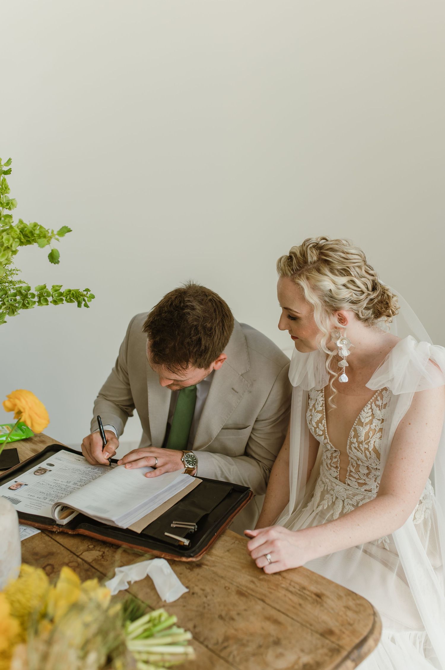 Couple signing wedding documents together at rustic wooden table with floral decorations.