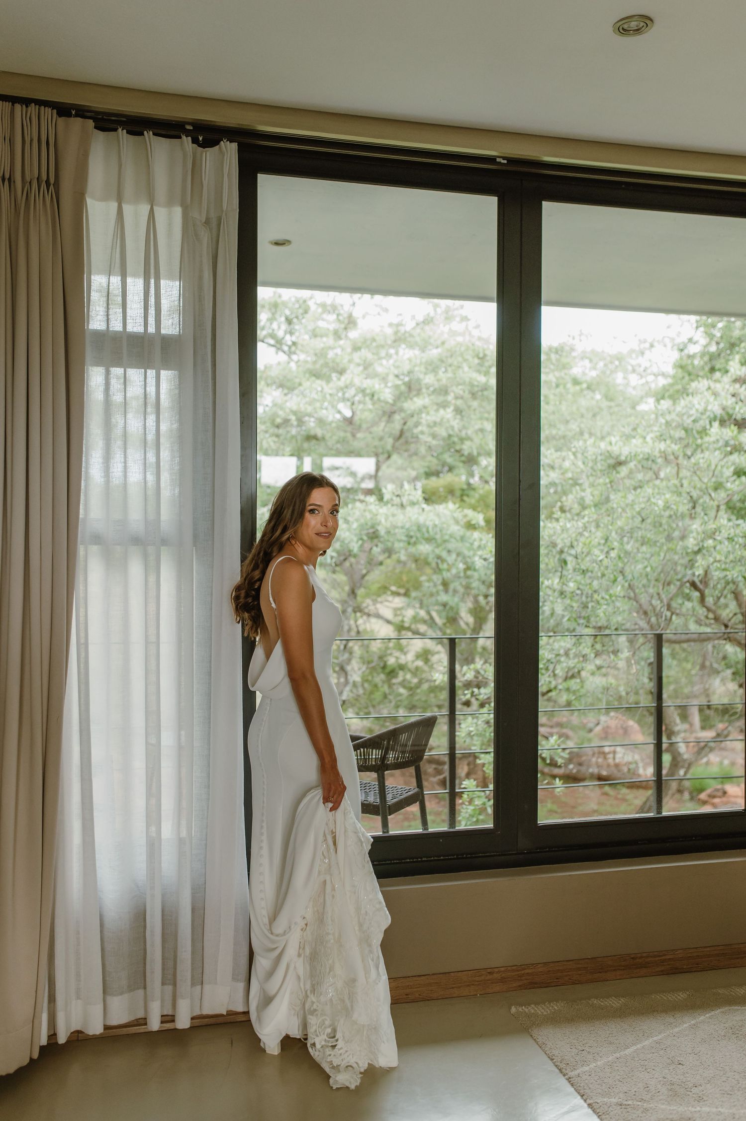 A flowing white dress moves elegantly against a backdrop of windows overlooking green trees.