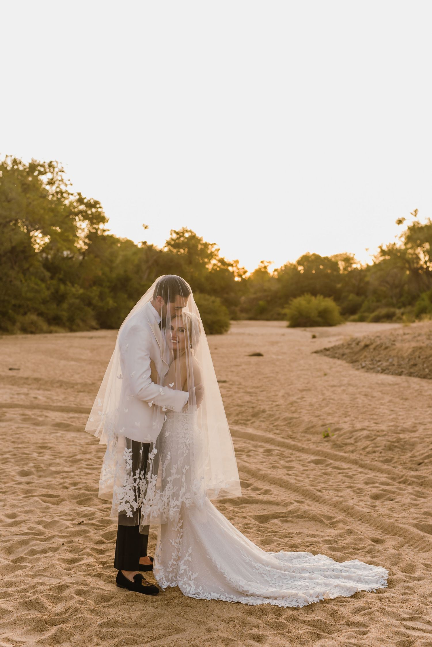 Romantic wedding couple poses on sandy beach at golden hour with flowing lace veil and dress.