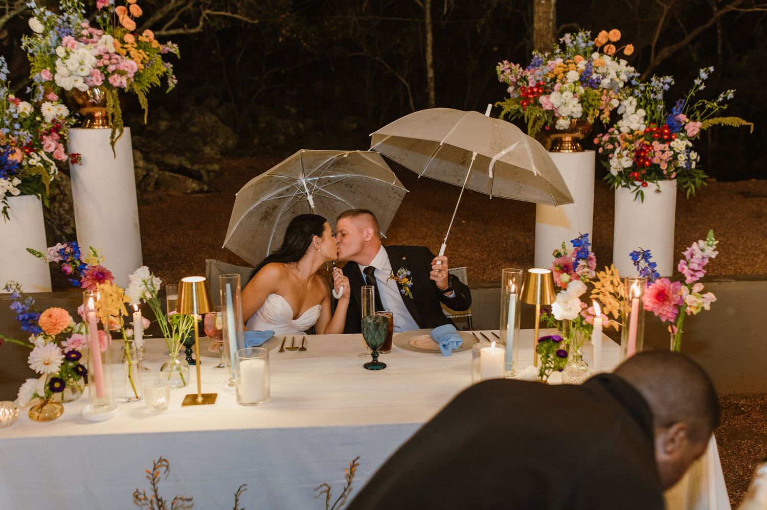 Bride and groom kissing under clear umbrellas at a candlelit sweetheart table with colorful floral decor.