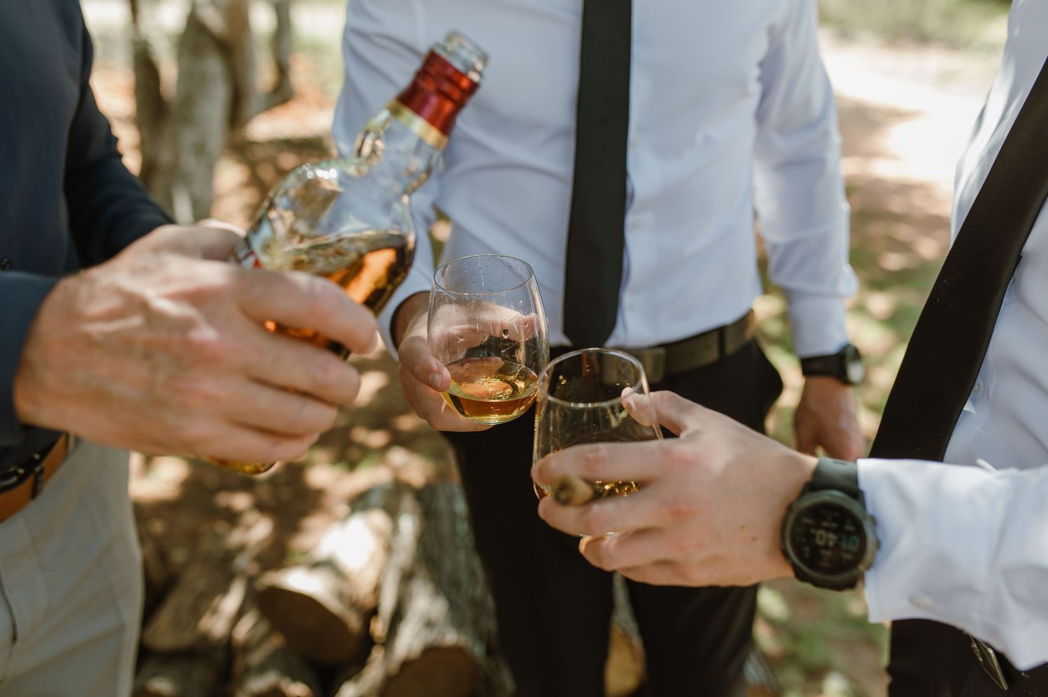 Groomsmen pouring whiskey into glasses outdoors before a wedding ceremony in a natural bush setting.