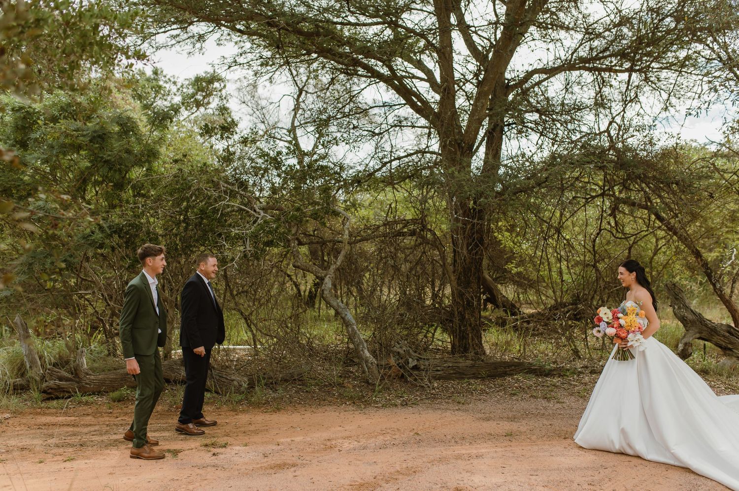 Bride in white gown holding colorful bouquet during first look moment with groom in nature.