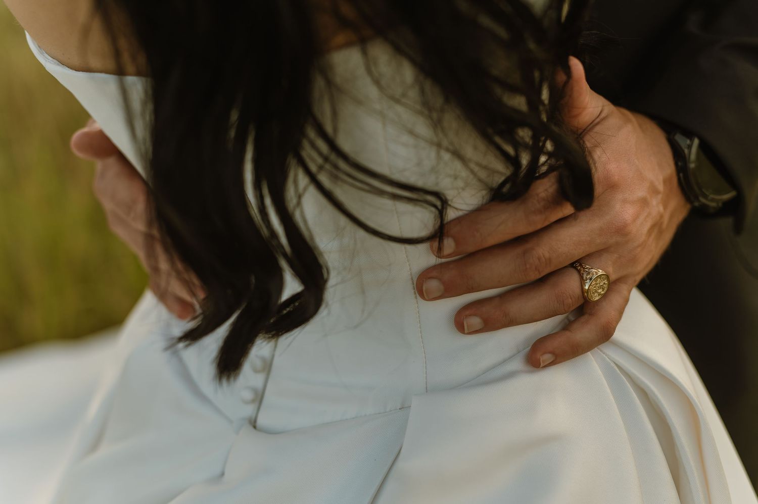 Close-up of grooms hand with gold ring resting on brides white wedding gown waist during portraits.
