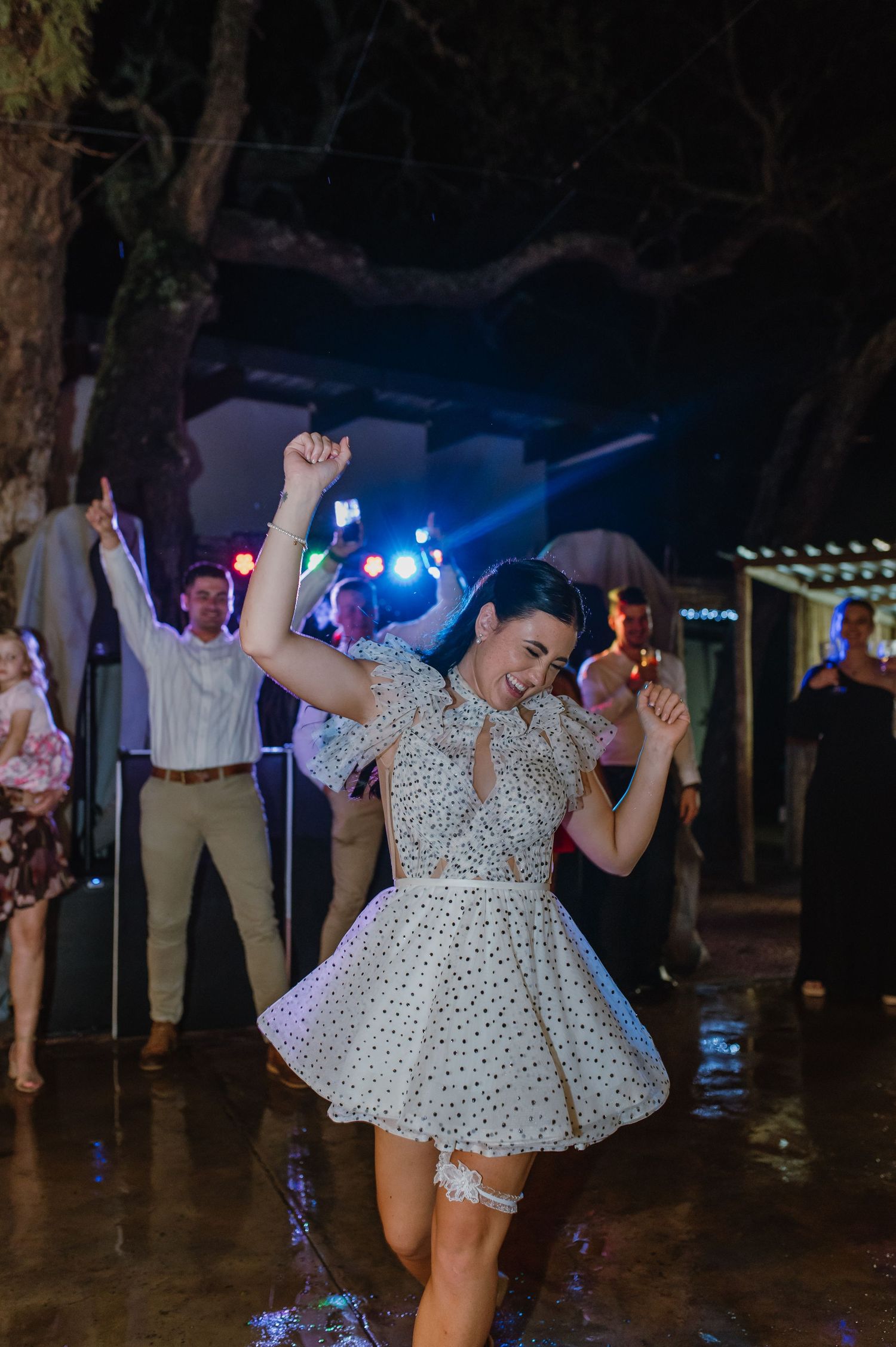 Bride in a white polka dot mini dress dancing joyfully on a wet outdoor dance floor at her wedding reception.