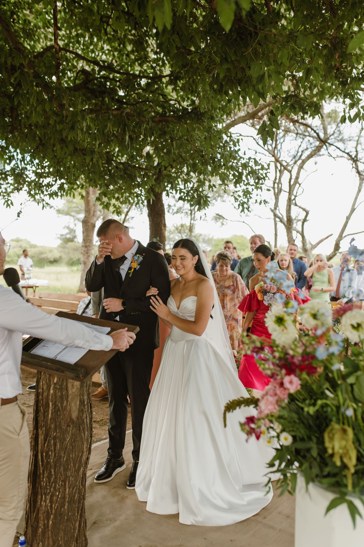 Emotional groom wiping tears beside bride in white gown during outdoor wedding ceremony.
