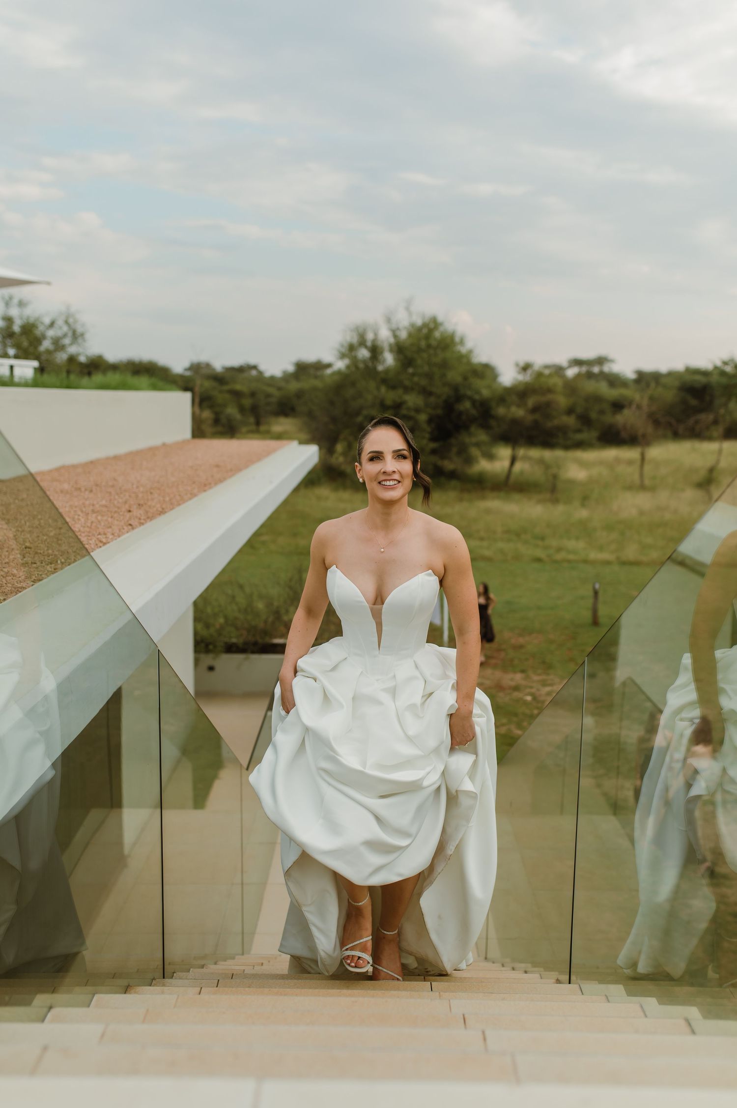 Bride in white dress poses on modern glass staircase with scenic outdoor backdrop.