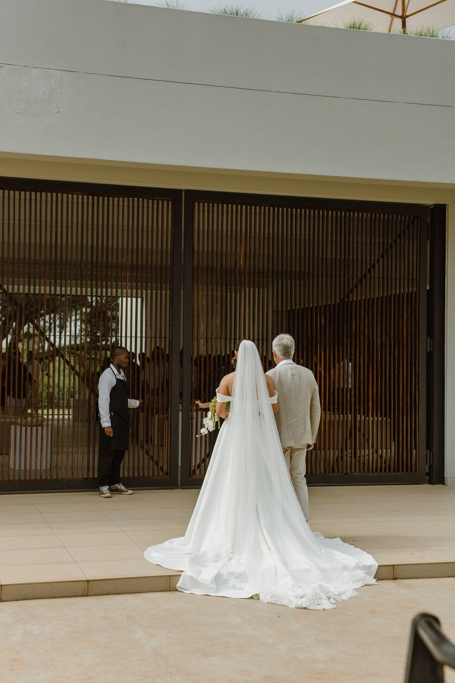 A bride and groom walk together through a modern architectural space with clean white walls and vertical slats.