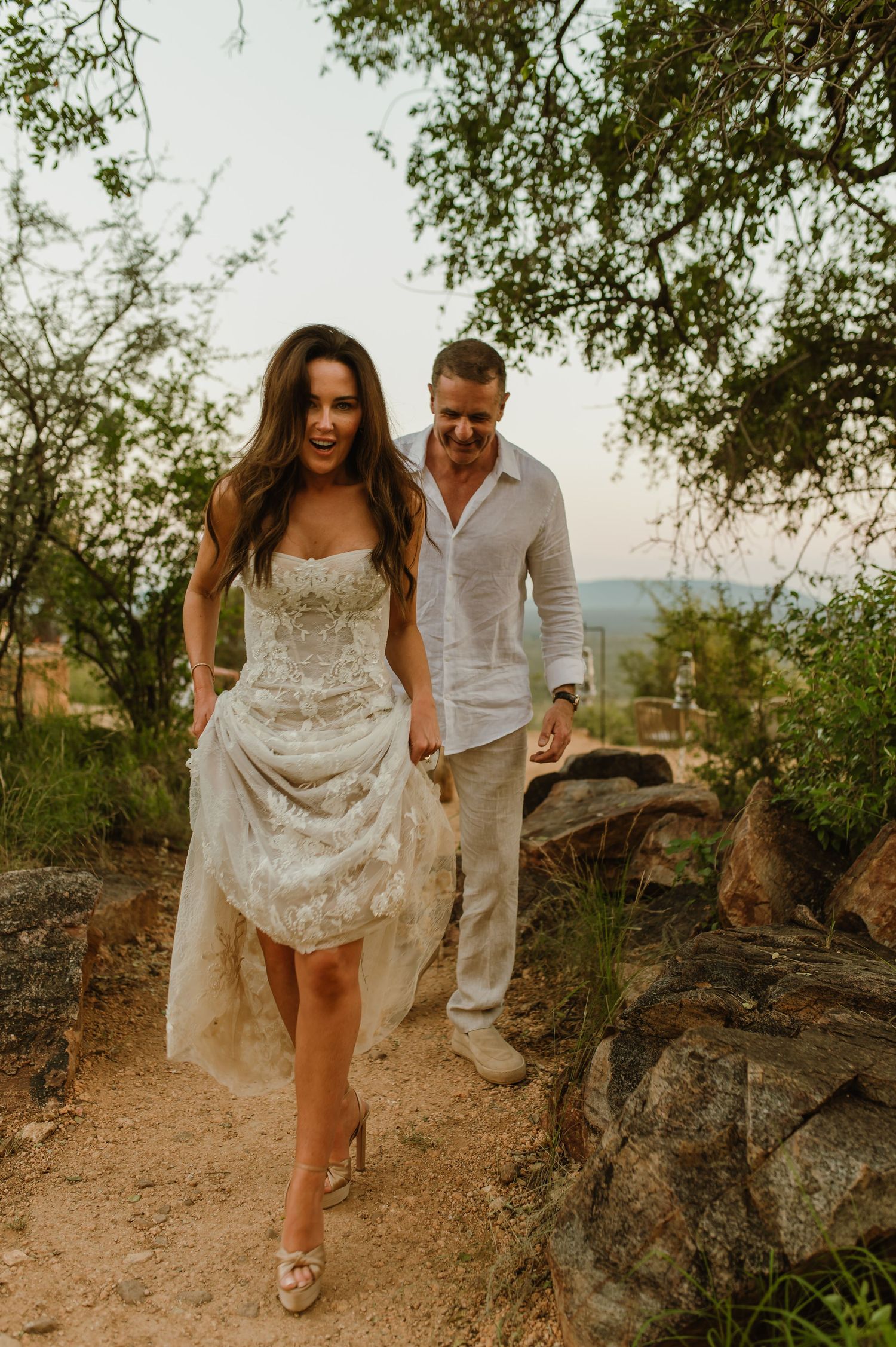 Laughing bride lifts lace wedding gown while walking rocky path with groom at sunset.