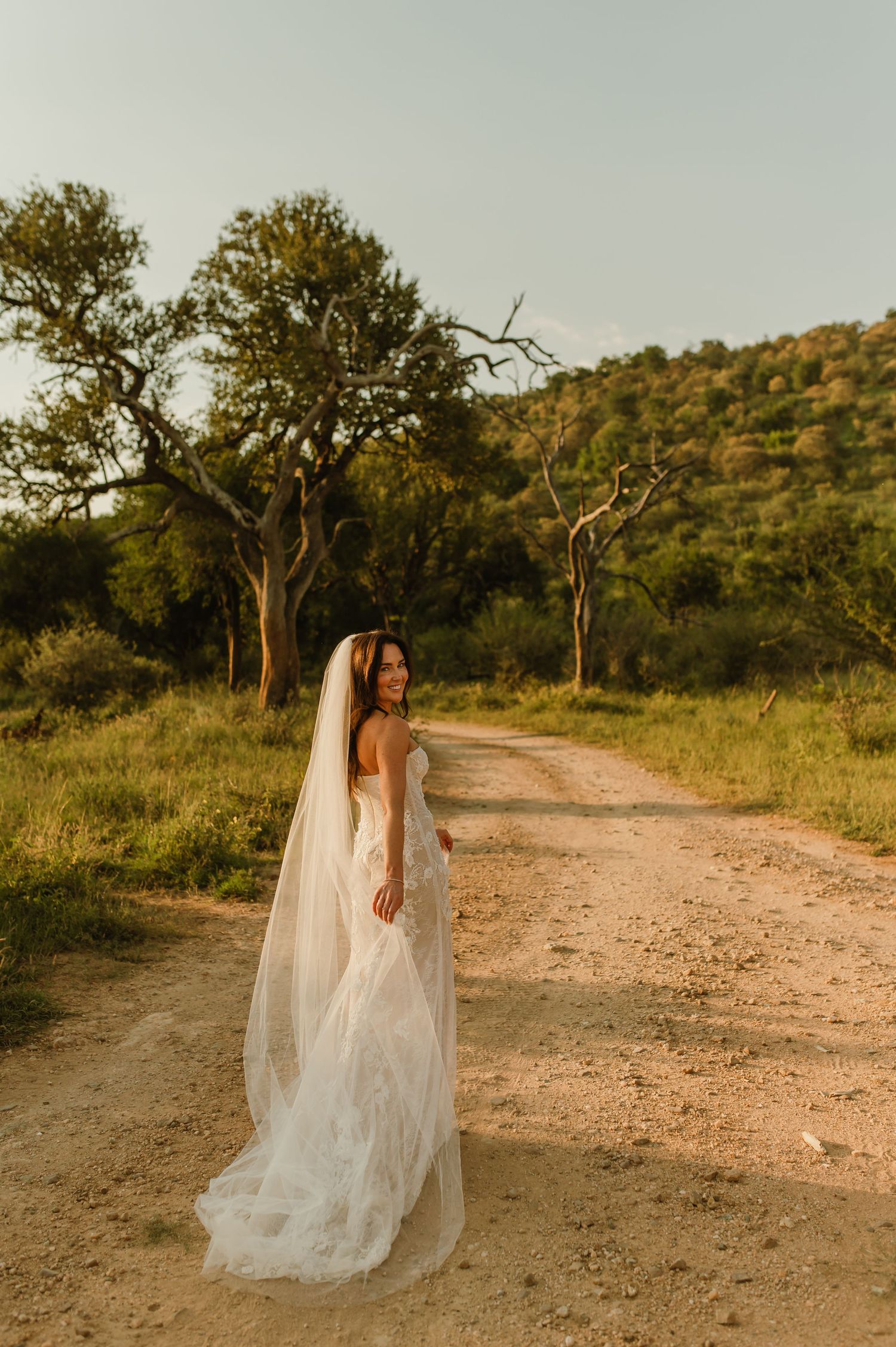 Bride in a flowing white gown and long veil stands on a sunlit dirt path surrounded by lush trees.