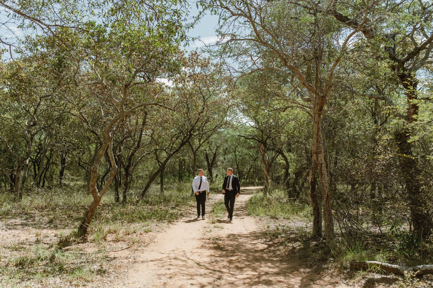 Two men in formal wedding attire walking together along a dirt path through a wooded forest.