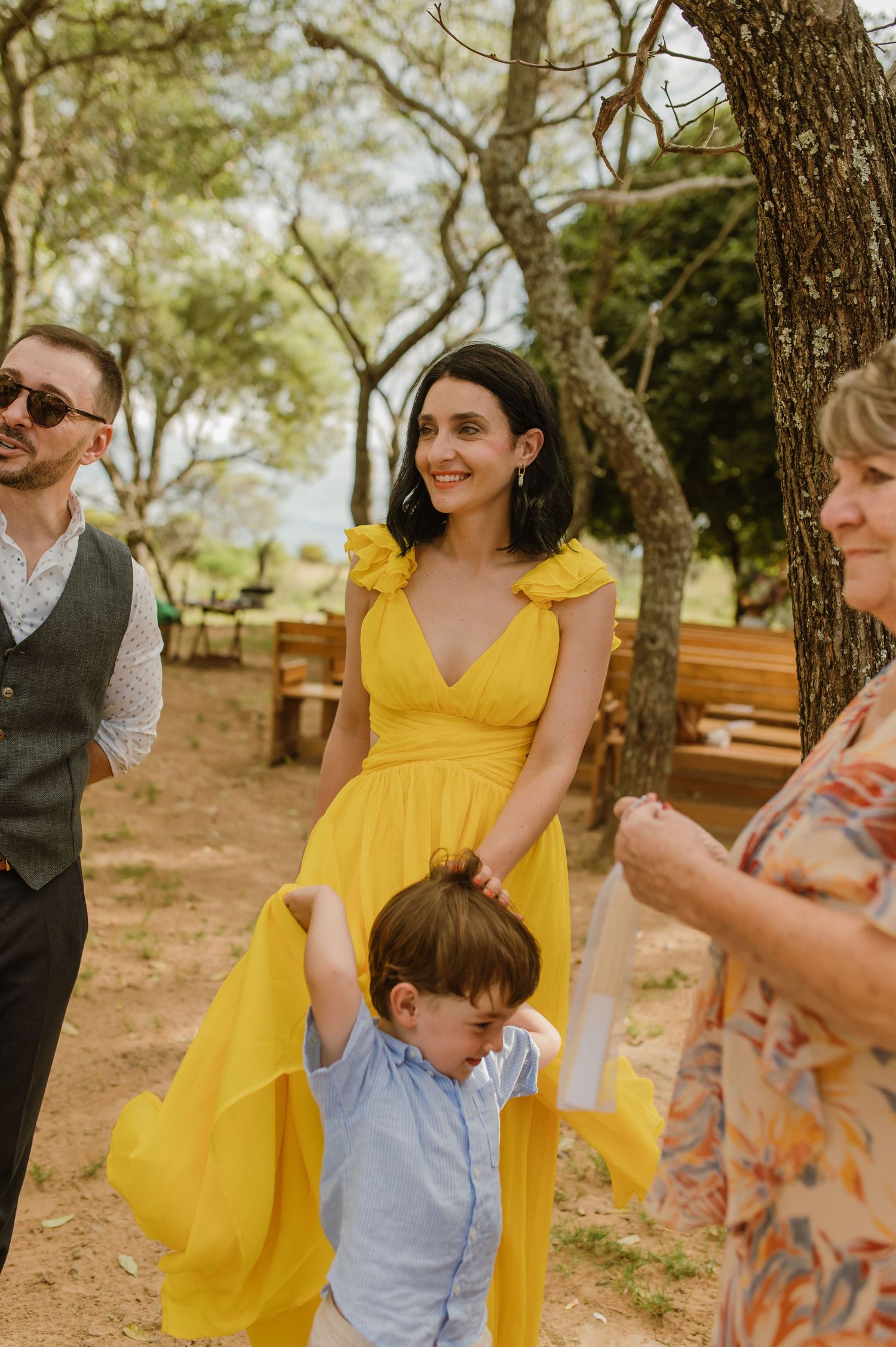 Woman in bright yellow dress holds fabric while young child grabs it at outdoor wedding celebration.