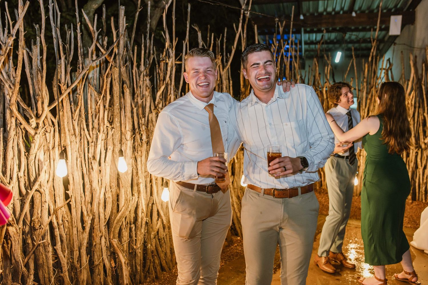 Two smiling male wedding guests pose with drinks near rustic branch installation at reception.