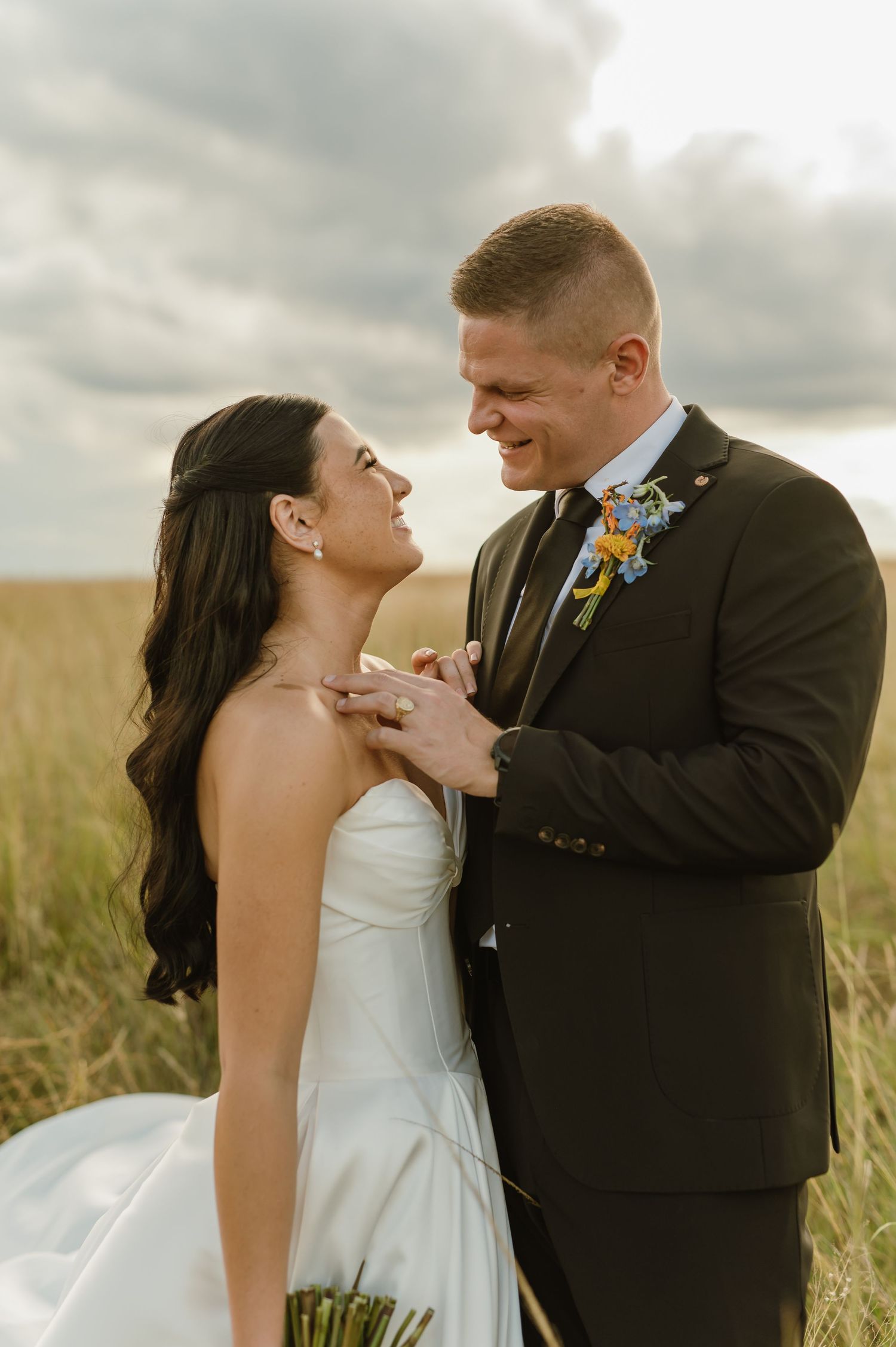 Bride and groom share a romantic moment in golden field, groom in dark suit with floral boutonniere.