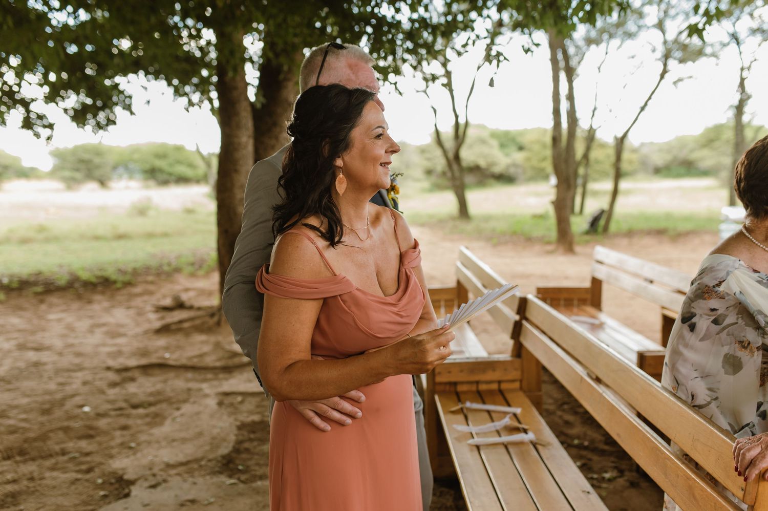 Smiling bridesmaid in pink off-shoulder dress stands outdoors at rustic wedding venue among trees.