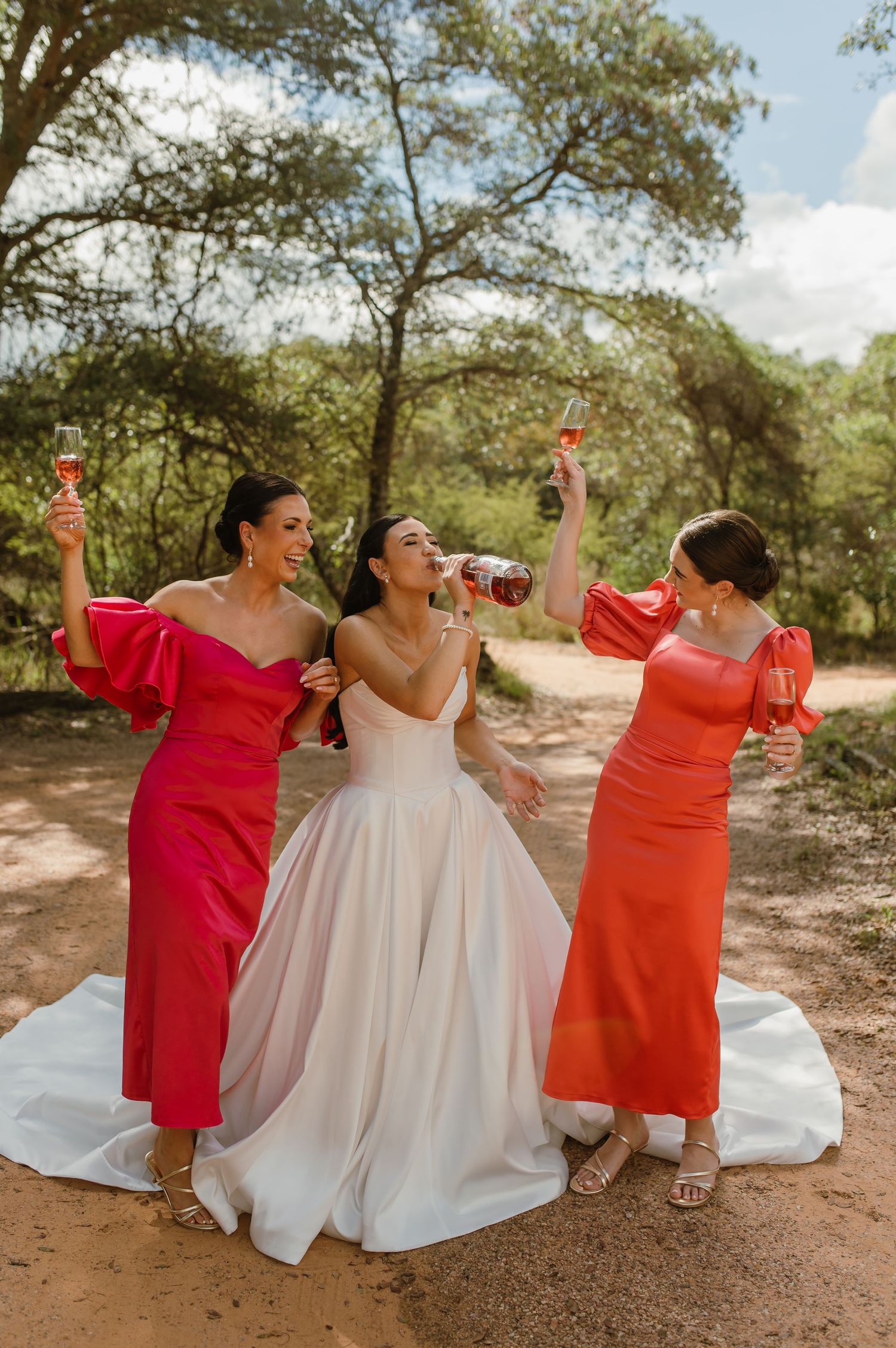 Bride in white ballgown toasting champagne with two bridesmaids in red and orange dresses outdoors.