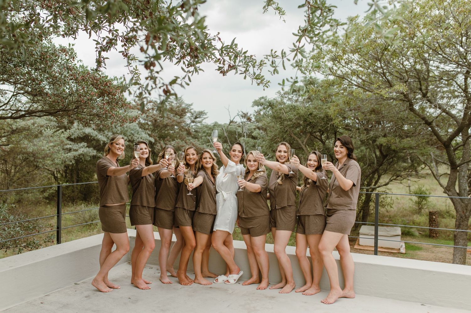 Wedding party in brown dresses and white gown posing outdoors under trees.