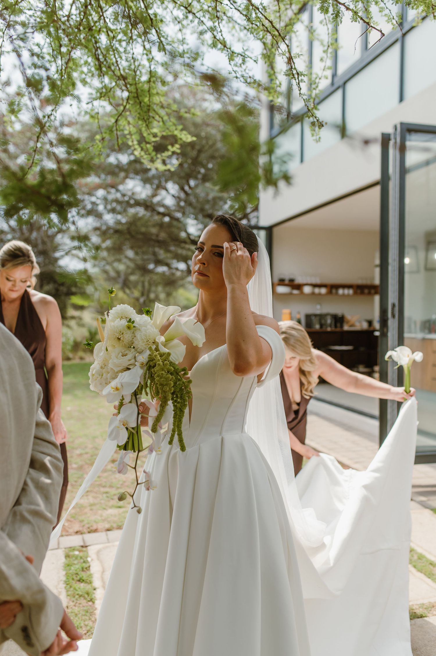 Wedding party outdoors with flowing white gown and orchid bouquet against modern architecture.