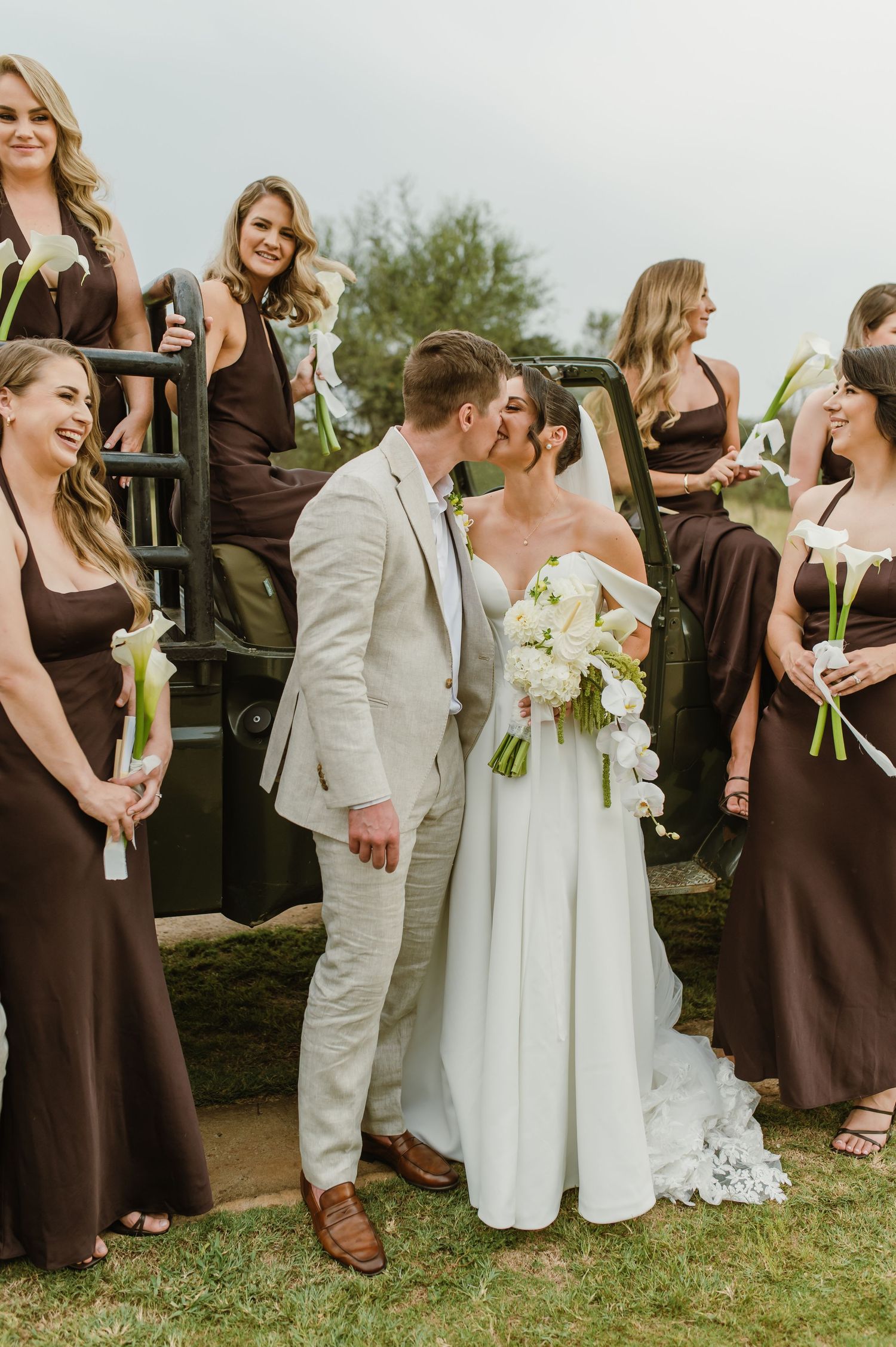 Wedding party wearing brown and white attire stands together outdoors on grassy area holding bouquets.