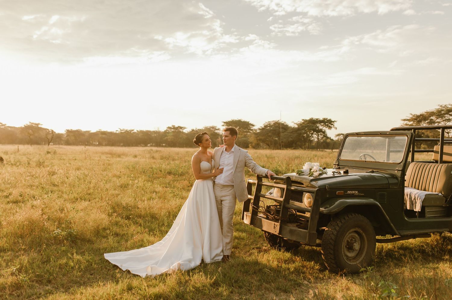 A romantic sunset wedding photo with a vintage Land Rover in a golden field.