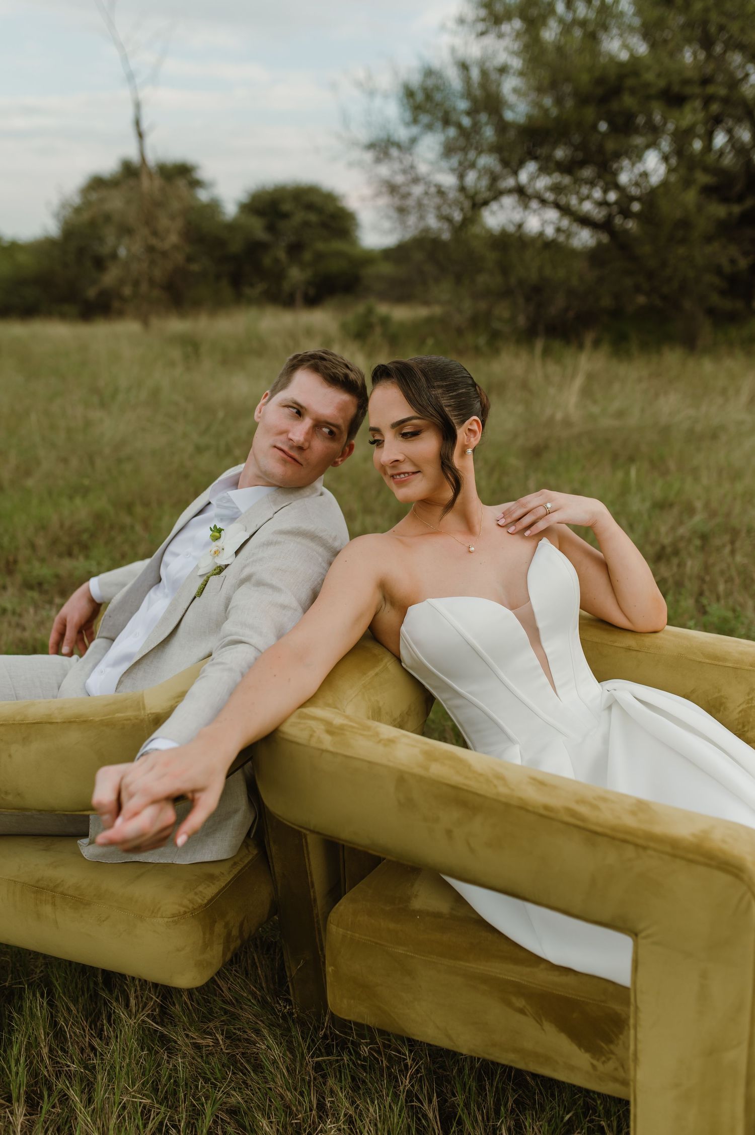 A couple in wedding attire relaxes on a yellow couch in a grassy field during a rustic outdoor photoshoot.