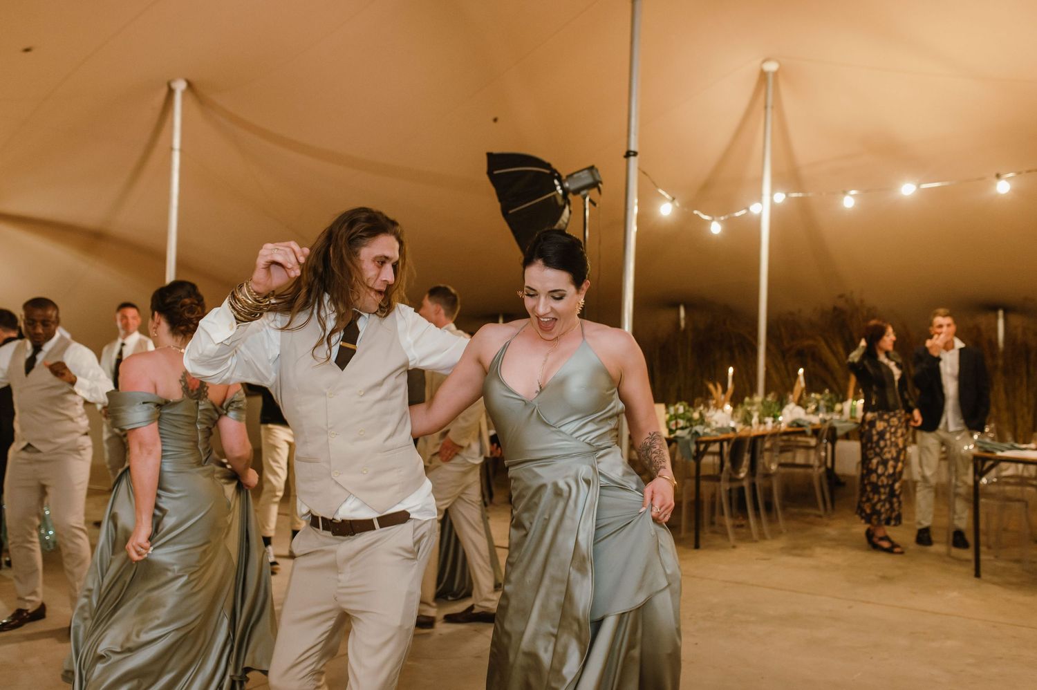Wedding party dancing in a tent venue with string lights and festive atmosphere.