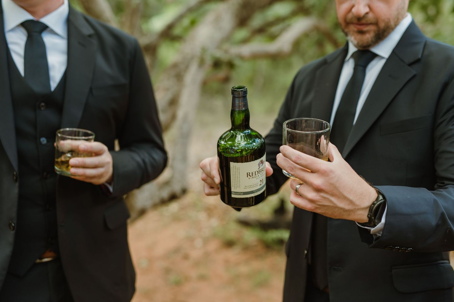 Wedding party members in black suits examining bottles in an outdoor setting.