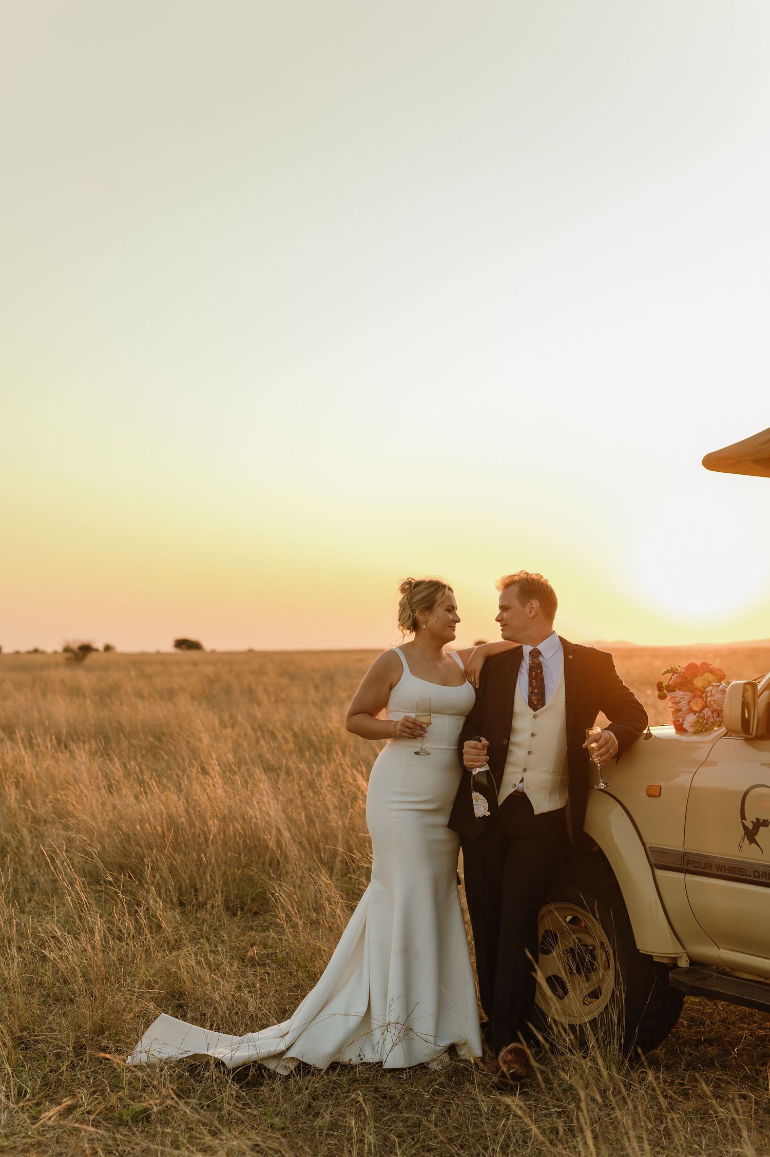 Newlyweds share intimate moment by vintage car in sunset field.