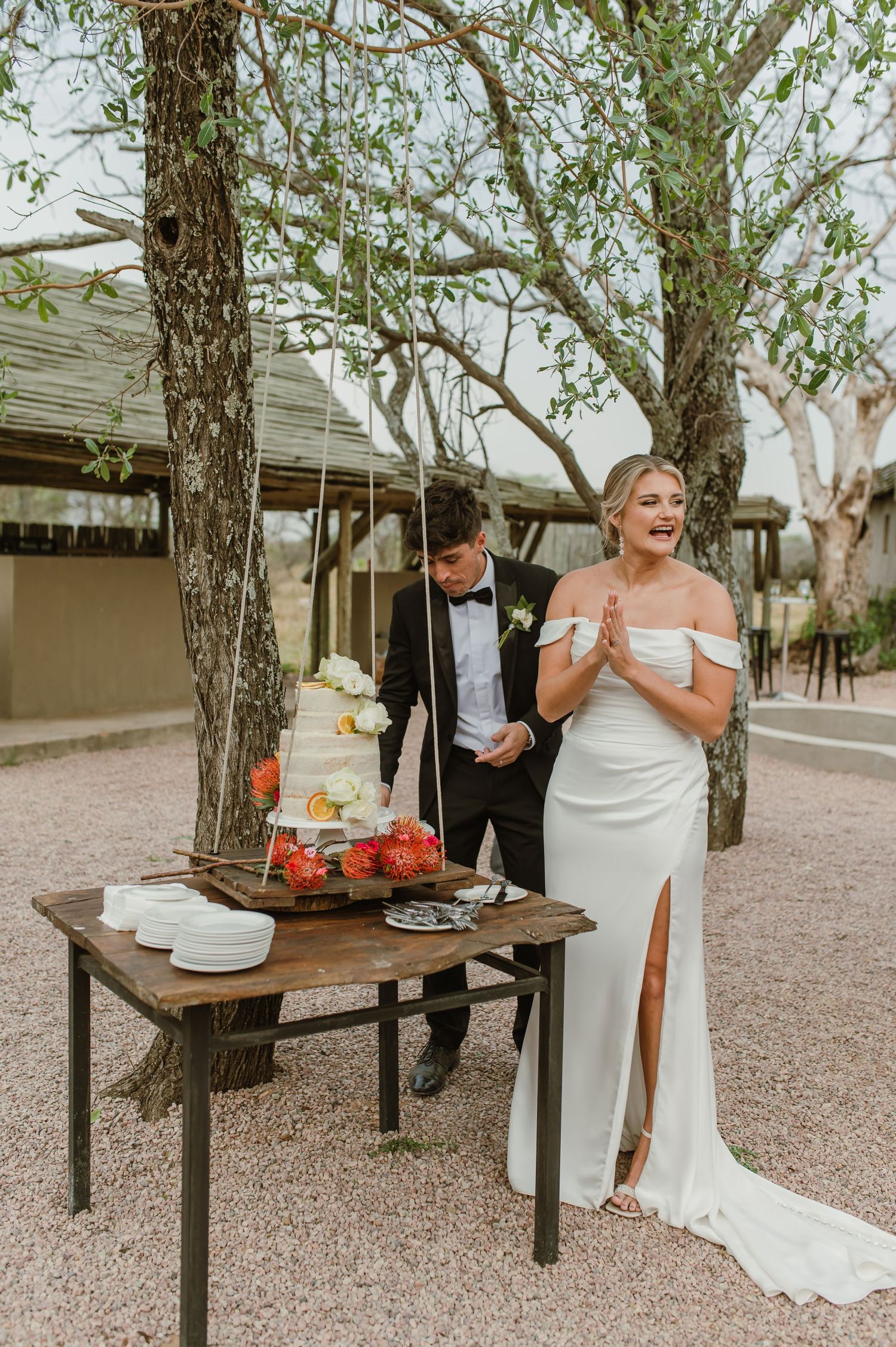 Couple laughing while sharing cake at their outdoor wedding reception with rustic wooden tables.