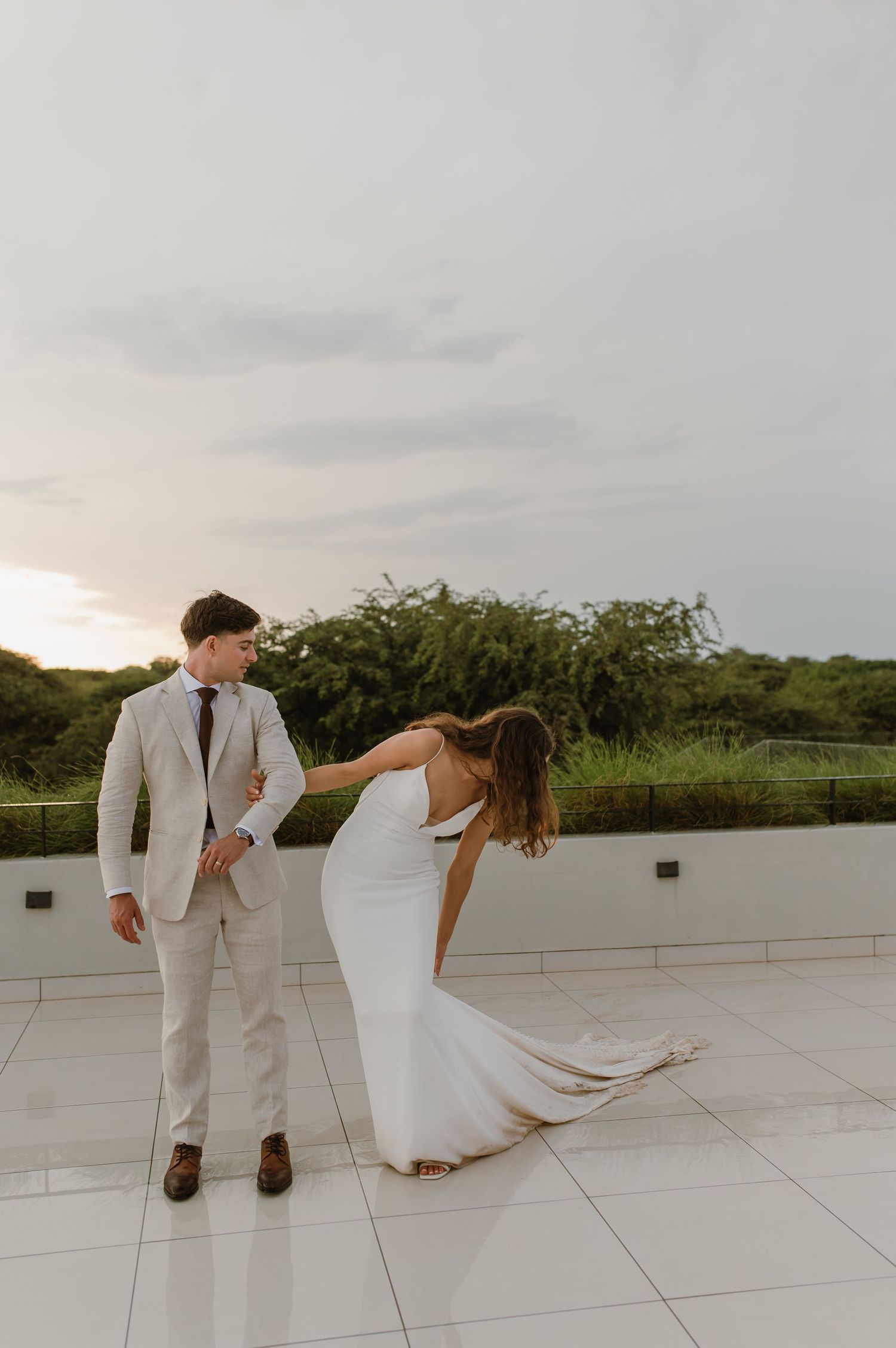 Playful wedding couple dances on white terrace with water view in background.