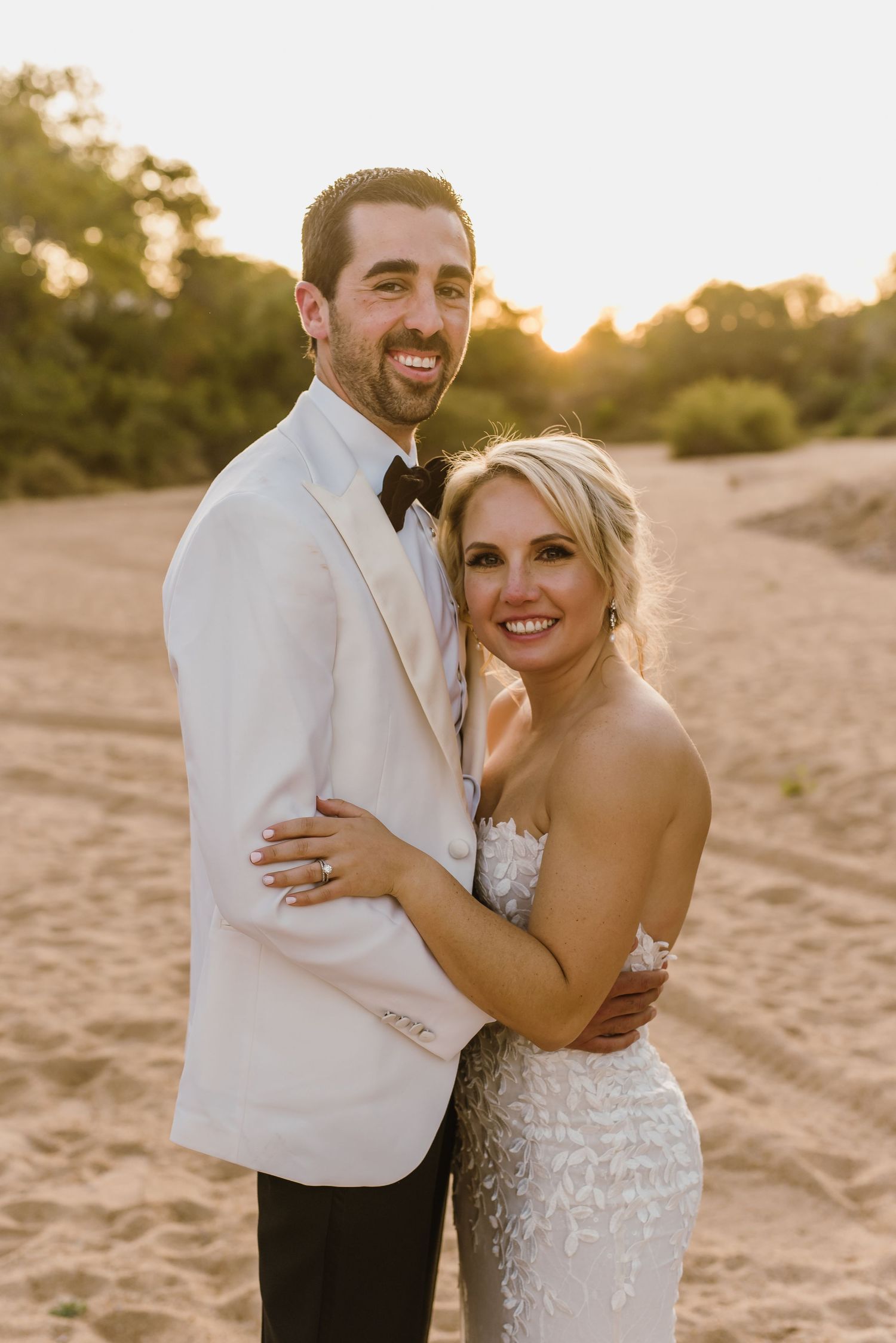 Joyful wedding couple laughs together on beach with sunset glow in background.