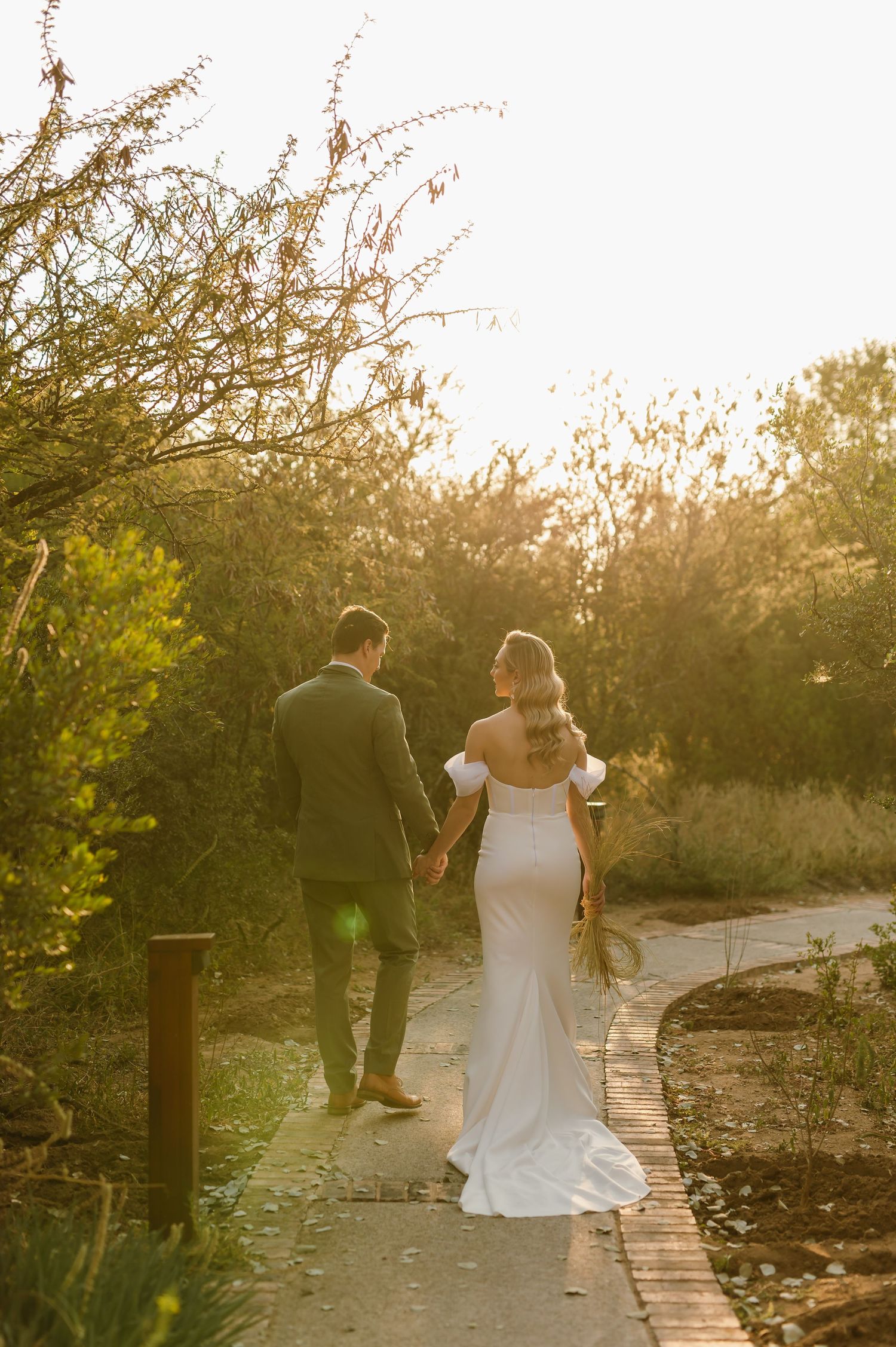 A couple walks along a scenic pathway during golden hour sunset with trees and natural scenery in the background.