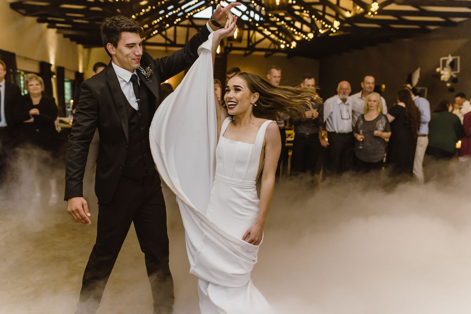 A couple shares their first dance in a romantic venue with exposed wooden beams and atmospheric fog effects.