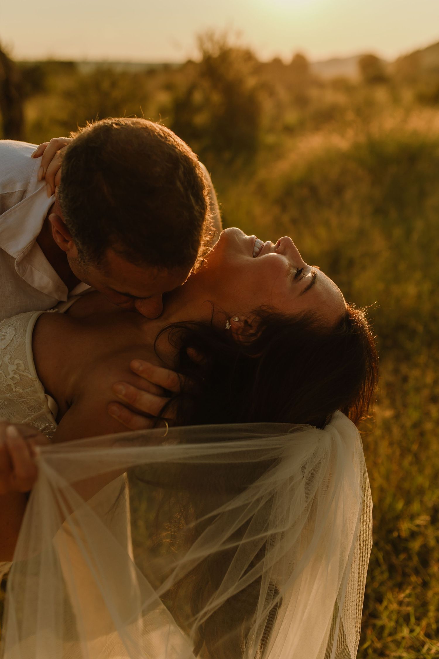 Groom kisses bride as she laughs joyfully in a sun-drenched field at golden hour.